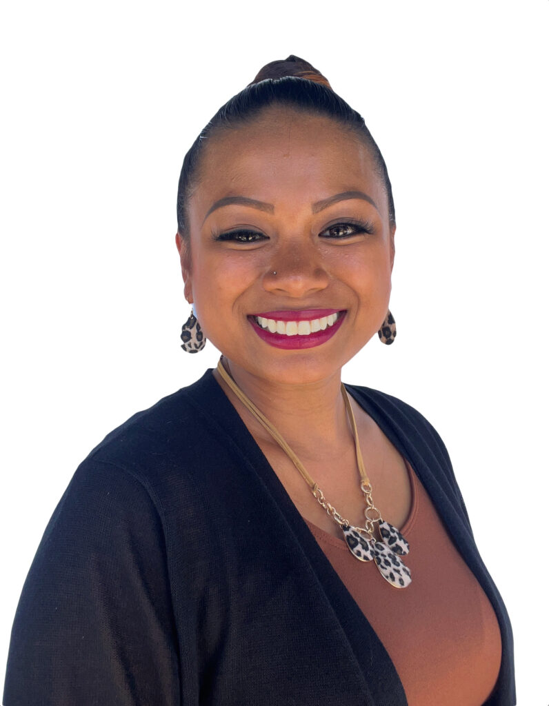 The image shows a woman smiling warmly against a white background. She has her hair pulled back in a neat bun and is wearing bold makeup, including eyeliner and deep pink lipstick. She is accessorized with matching leopard-print earrings and a necklace. Her outfit consists of a black cardigan over a brown top. Her expression is friendly and inviting, and the overall appearance gives off a confident and professional vibe.