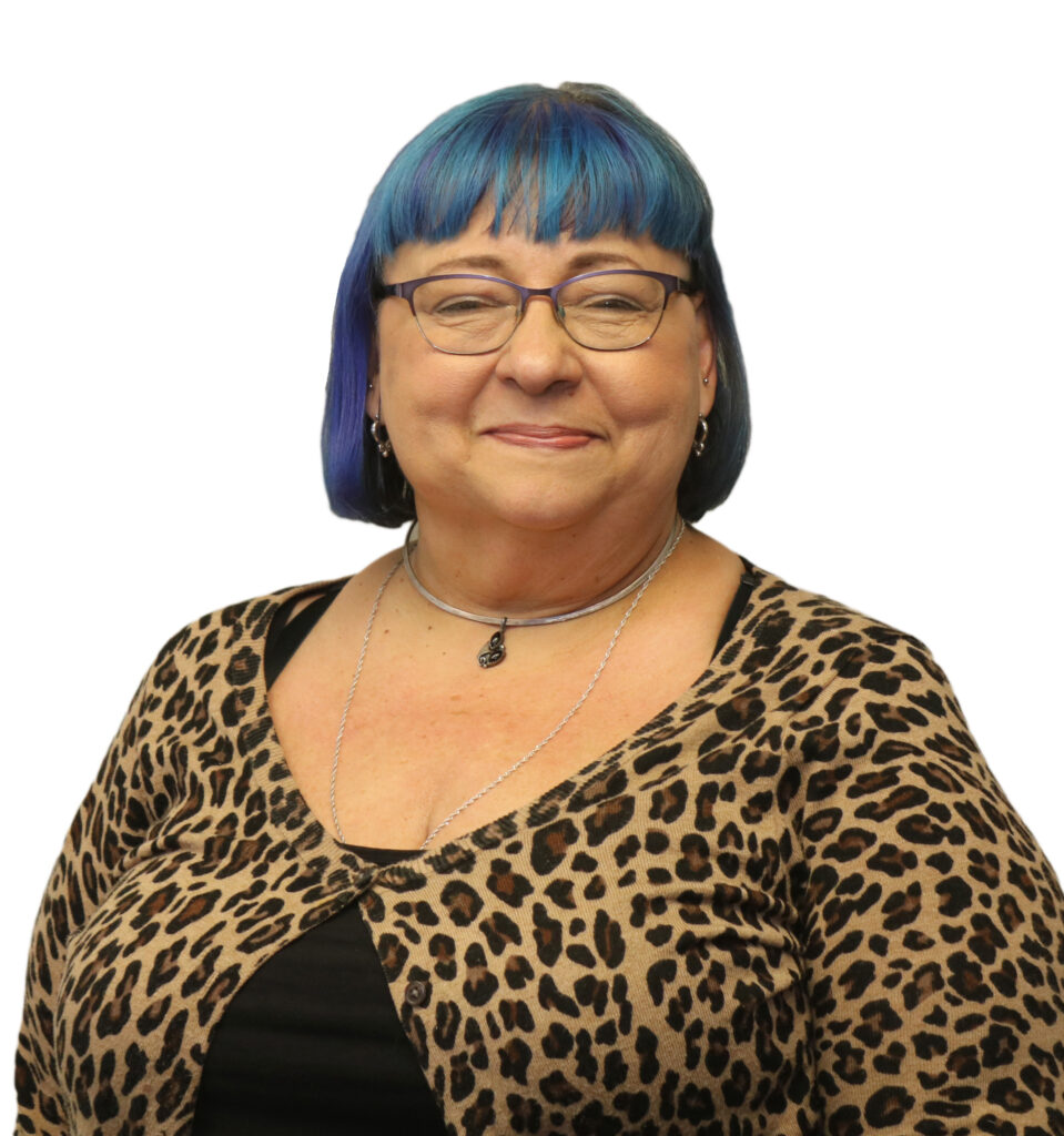 The image shows a woman smiling softly in front of a white background. She has short hair with a vibrant blue color, styled with bangs. She is wearing glasses and accessorizes with a silver choker necklace and a longer necklace with a black pendant. Her outfit includes a leopard-print cardigan over a black top, adding a bold, stylish touch to her look. Her expression is warm and calm, giving off a confident and approachable vibe. The overall tone is professional with a unique sense of personal style.