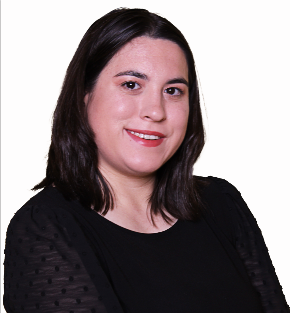 The image shows a woman smiling against a plain white background. She has shoulder-length dark hair, parted in the middle, and is wearing subtle makeup with a natural pink lip color. She is dressed in a black top with sheer sleeves that have textured dots, adding a stylish touch. Her expression is friendly and approachable, and the overall image has a professional, yet relaxed, tone, likely intended for a formal or business-related portrait.