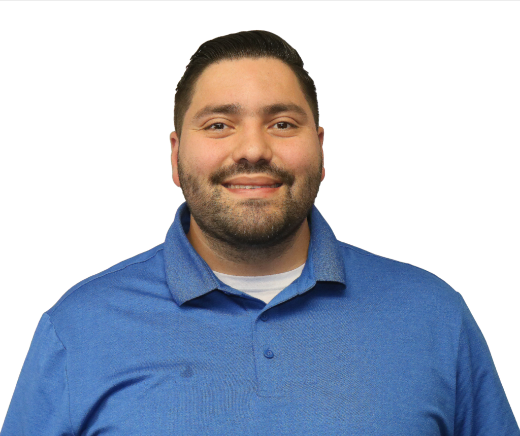 The image shows a man smiling in front of a plain white background. He has short, neatly styled dark hair and a well-groomed beard. He is wearing a blue polo shirt with a collared neck, and underneath, a white t-shirt is slightly visible. His expression is friendly and approachable, and the image is a professional business portrait.