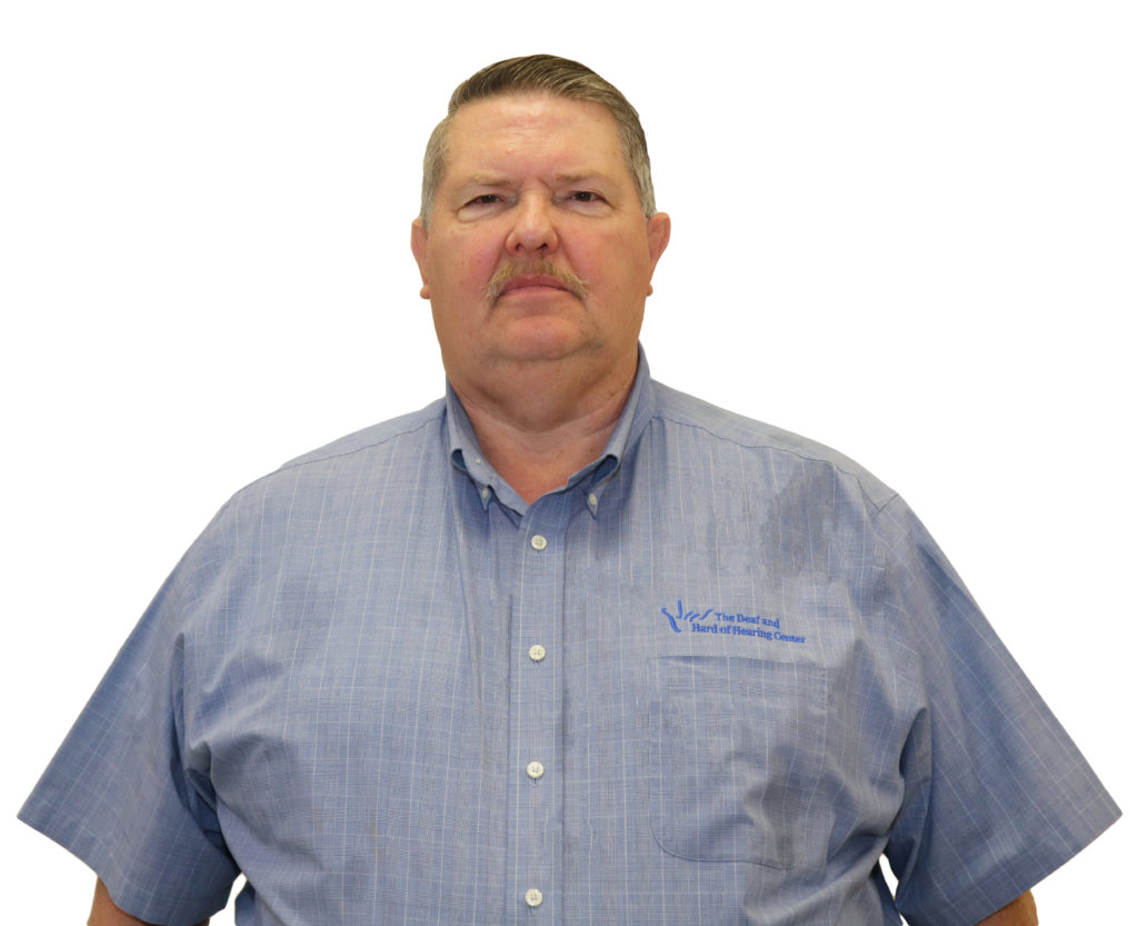 The image shows a man standing against a white background with a neutral expression. He has short, neatly combed gray hair and a mustache. He is wearing a light blue, short-sleeved button-up shirt with a subtle checkered pattern. The shirt has an embroidered logo on the left side that reads “The Deaf and Hard of Hearing Center.” His posture is formal, and the overall image presents a professional appearance.